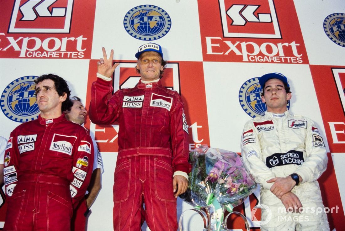 Niki Lauda, recién coronado Campeón del Mundo al terminar en 2ª posición, sube a lo más alto del podio mostrando tres dedos por cada título junto a Alain Prost, 1ª posición, y Ayrton Senna, 3ª posición.